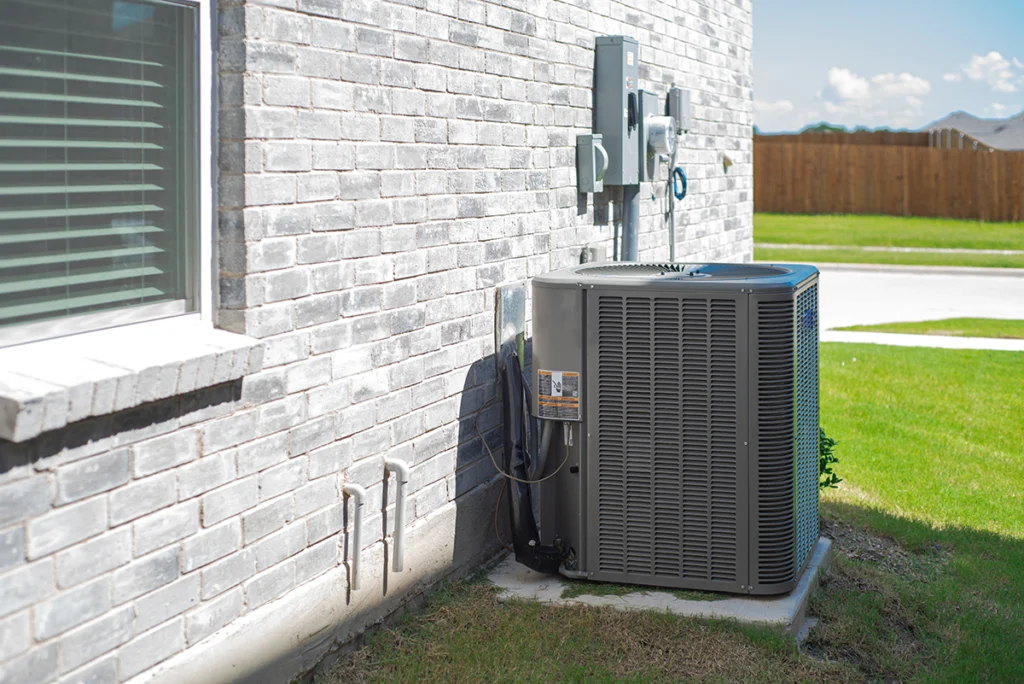 Residential outdoor AC condenser unit installed beside a brick home with visible electrical and refrigerant connections. Best for blogs about air conditioning systems, AC replacement, condenser units, and residential cooling equipment.